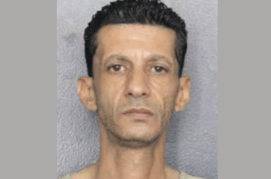Portrait-style booking photo of an adult male with a serious expression, wearing a beige shirt in front of a neutral backdrop.