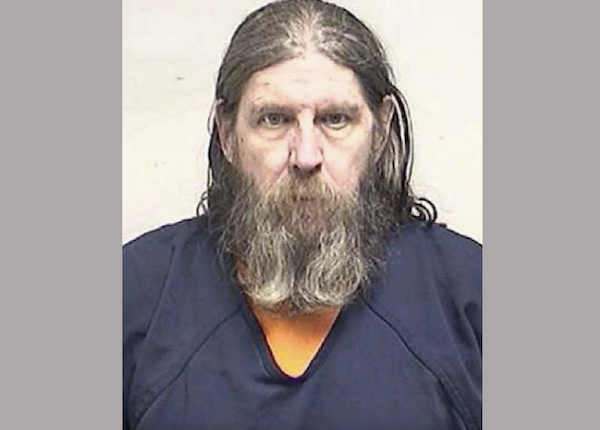 Police booking photo of a man with shoulder-length hair and a full beard, wearing an orange shirt under a navy blue jail garment.