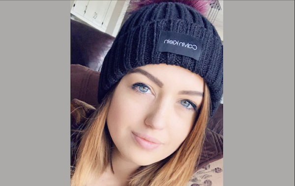 Young woman with blue eyes and straight hair poses for a photo in a room, dressed in a dark winter hat featuring a visible Calvin Klein label.