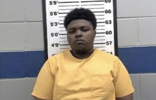 A young person in a yellow inmate uniform is photographed against a gray and blue wall with a measuring chart in a mugshot setting.