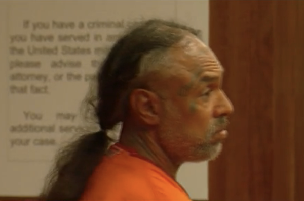 Side profile of an individual in an orange jail uniform during a court hearing, with a sign visible in the background.