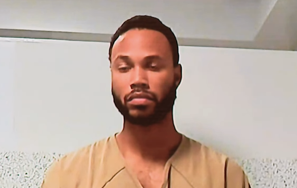 Individual wearing a beige prison jumpsuit appears behind glass in a holding area with white walls.