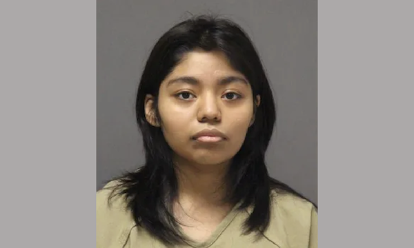 Straight-on portrait of a woman with black hair, dressed in a beige top, looking directly at the camera with a blank expression, gray backdrop behind her.