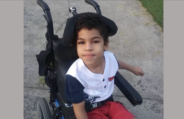 Child in a wheelchair outdoors, dressed in a white and blue top with red pants, poses on a concrete path next to grass, appearing calm and content.