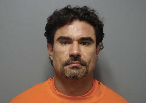 Mugshot of a middle-aged man with tan skin and facial hair, dressed in an orange garment, standing before a plain gray background.