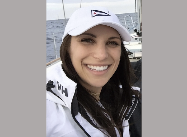 Woman with straight brown hair and white sailing gear takes a selfie on a boat, with the sea and cloudy sky behind her.