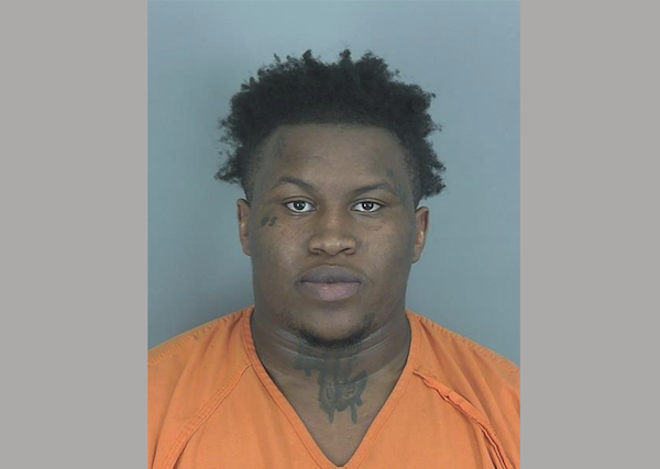 A person in an orange jail uniform is photographed head-on for a mugshot, with a neutral blue-gray backdrop.