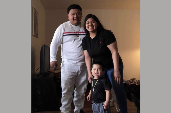 Family portrait shows an adult male in white, an adult female in black, and a small boy posing together inside a cozy home.