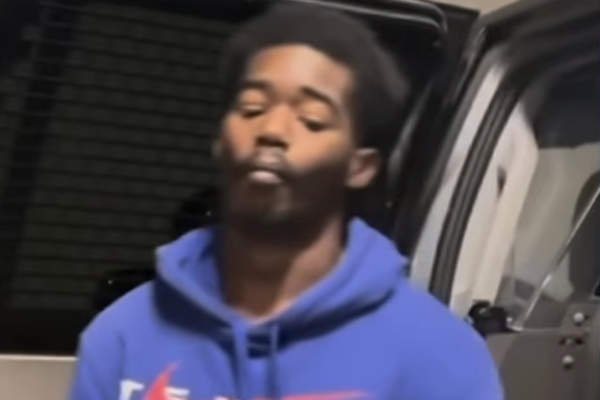 Young man wearing a blue sweatshirt is shown from the shoulders up, standing slightly off-center in front of a dark window.