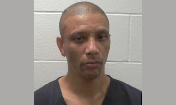 A man in a black top photographed in front of a white brick wall, facing forward with a serious expression.