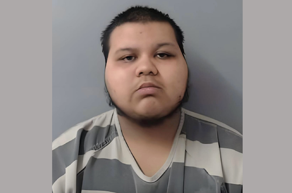 Mugshot of an individual with short dark hair and a serious face, dressed in a standard-issue prison jumpsuit, positioned before a plain wall.