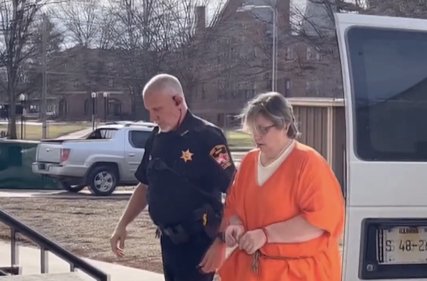 A law enforcement officer walks alongside a handcuffed woman dressed in orange, outside near a courthouse parking lot on a sunny day.