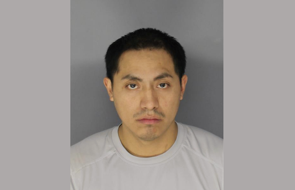 Police booking photo showing a man with medium complexion, dark eyes, short hair, and a serious expression, wearing a pale gray athletic shirt.