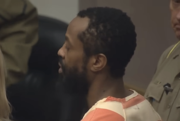 Side profile of a man dressed in a prison jumpsuit during a court appearance, his expression neutral.