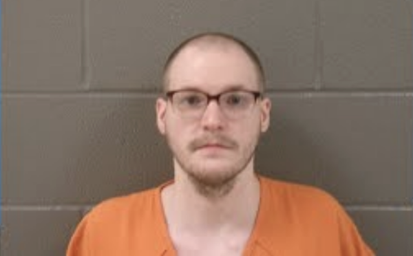 A serious-looking male individual in an orange inmate uniform, standing in front of a neutral, gray brick background.