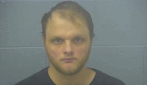Front-facing mugshot of a young adult male with thinning hair, a trimmed beard, and a neutral expression, set against a blue-gray jail background.