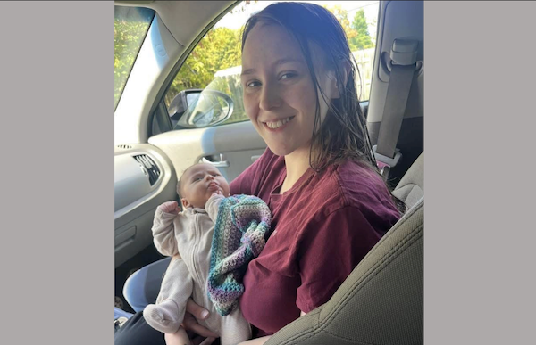Inside a vehicle, a woman wearing a maroon shirt cradles an infant dressed in a pale outfit, both facing the camera.