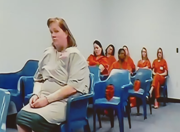 Several individuals dressed in orange and one in gray, all seated in rows of blue chairs inside a sparse room, suggesting a correctional facility or courtroom setting.