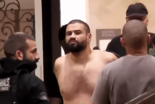 Police officers stand around a bearded, shirtless man in handcuffs, who appears to be in custody.