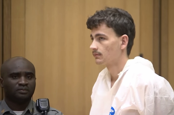 Side profile of a man dressed in a white forensic jumpsuit standing in a courtroom, appearing serious and subdued during legal proceedings.