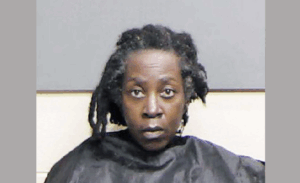 Mugshot of a woman with tightly curled hair and a serious expression, dressed in standard jail attire, seated in front of a plain wall.