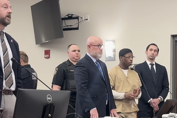 Five men stand at the front of a courtroom, including a defendant in beige jail attire, two men in suits, a police officer, and another man gesturing while speaking.