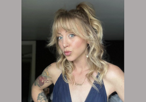 Young woman with curly blonde hair and bangs, wearing hoop earrings and a necklace, stands with her arm bent, revealing colorful tattoos on her upper arm.