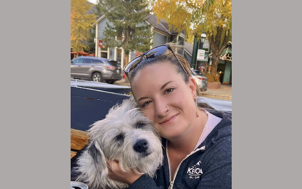 Woman with sunglasses resting on her head poses affectionately with a shaggy dog, sitting outside near a street lined with fall foliage.