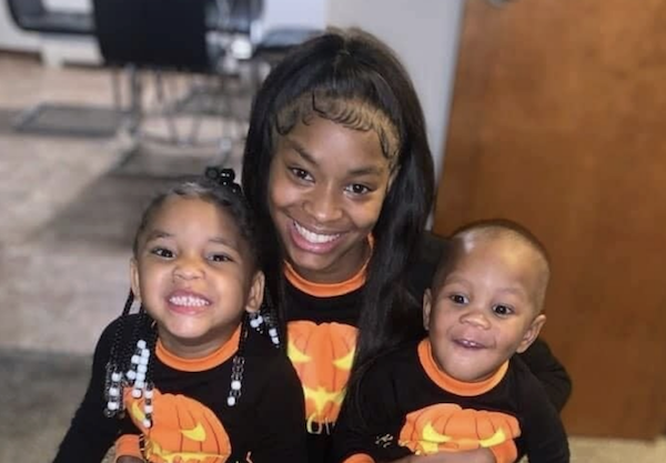 Mother and her two children pose together and grin, dressed in coordinated black and orange pumpkin outfits, in a cozy home setting.