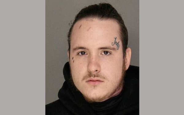 Portrait-style police booking photo showing an adult male with facial tattoos, a mustache, and slight beard, dressed in a dark garment, with a neutral gray backdrop.