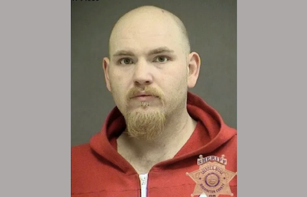 Mugshot of a light-skinned man with light facial hair and a serious expression, dressed in a red jacket, with a law enforcement badge overlayed on the image.