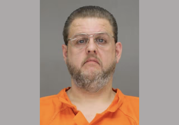 Mugshot of a man with a short beard, slicked-back hair, and clear eyeglasses, dressed in an orange prison jumpsuit.