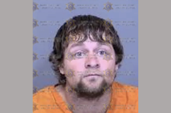 Booking photo showing a male with light skin, curly hair, and facial hair, dressed in orange jail clothing, with official watermark text across the image.
