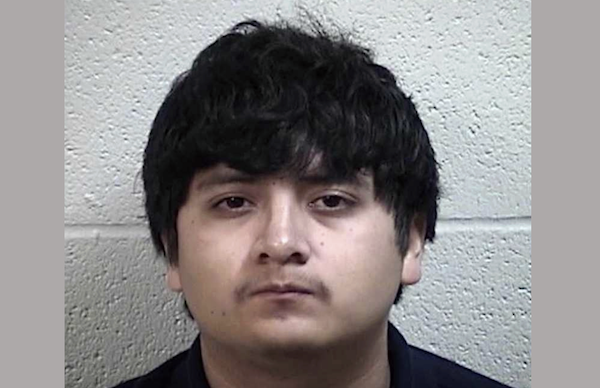 Mugshot of a male with medium-length black hair and light skin, wearing a dark shirt, with a textured, pale wall as the background.
