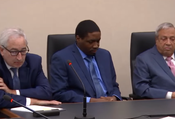 A man in a blue suit sits between two other men at a courtroom bench, with microphones visible in front of them.