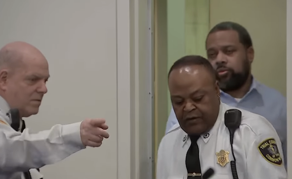 Law enforcement officers lead a handcuffed individual through a doorway during court proceedings.