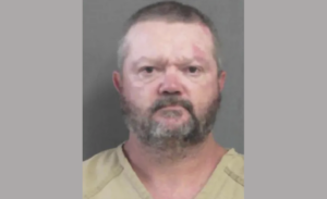 Portrait-style police booking photo showing a stern-faced man with facial hair and visible skin blemishes, wearing a tan jail outfit.