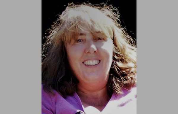 Portrait of a woman outdoors, with sun-lightened hair and a cheerful expression, dressed in a pink shirt.