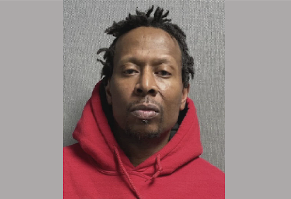 Portrait of a man in a red hoodie against a plain gray wall, gazing straight ahead with a serious look.