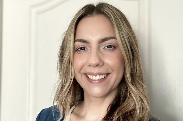 Smiling woman with light brown hair and fair skin wearing a dark blue top, posing indoors against a neutral background.