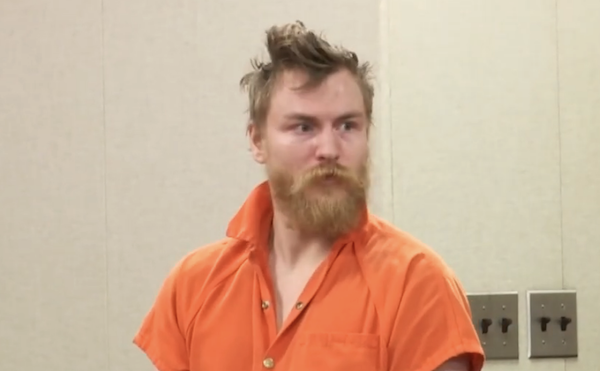 Individual dressed in an orange prison uniform, with a stern expression and unkempt hair, sits in a neutral-colored room.