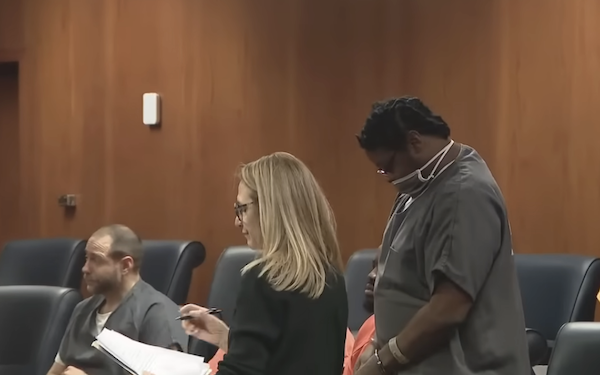A group of defendants in jail attire sit and stand during a court proceeding, as a woman with long blonde hair moves past them, set against a background of wooden paneling and blue chairs.