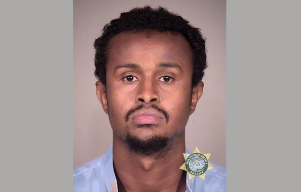 Mugshot portrait of an adult male with dark hair and facial hair, set against a plain background, featuring an official emblem near his shoulder.