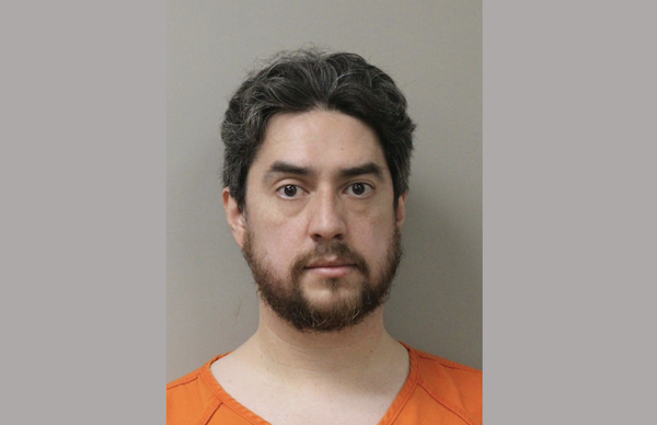 Booking photo showing a bearded male with medium-length black hair, dressed in an orange jail uniform.