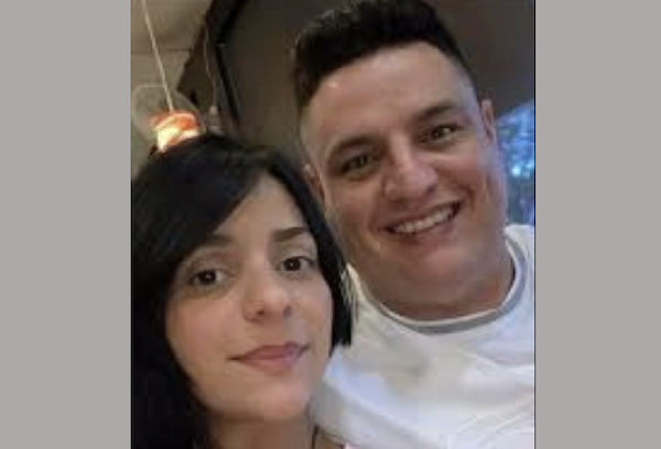 Two people pose for a selfie, with the woman in the foreground wearing a pale pink blouse and the man behind her in a white shirt, both looking directly at the camera and smiling.