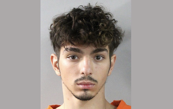 A head-and-shoulders image of a male with olive skin, short curly hair, and a faint mustache and goatee, dressed in an orange top, looking directly at the camera.