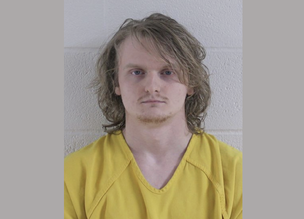 Mugshot of a man with pale skin, light-colored eyes, and disheveled hair, dressed in a yellow jail uniform, posing against a textured wall.
