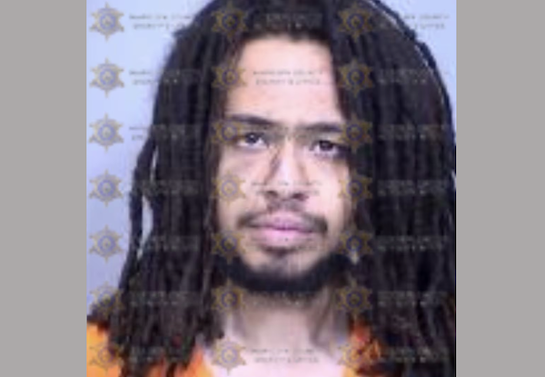Booking photo showing a young adult male with dark dreadlocks, facial hair, and an orange jumpsuit, set against a neutral background marked with law enforcement insignias.