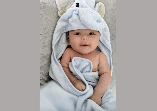 Infant wearing an elephant-themed hooded towel, looking up and partially wrapped, resting on a plush surface.