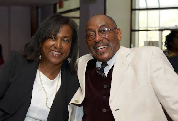 Two people pose together for a photo, the woman in a black blazer and pearl earrings, the man in a cream jacket and maroon vest, both looking at the camera and smiling warmly.